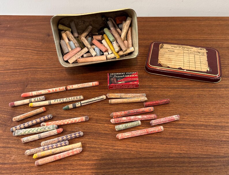 May include: An assortment of vintage crayons in various colors, scattered on a wooden surface. A metal tin and a small box of absorbent filters are also present. Some crayons are labeled with the words "Crayon" and "Visualizer".