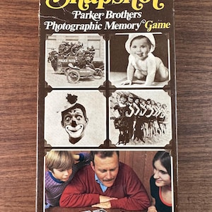 May include: Vintage "Snapshot" Parker Brothers Photographic Memory Game box. The box features a collage of black and white images, including a baby, a clown, and a group of people. The game is being played by a family.