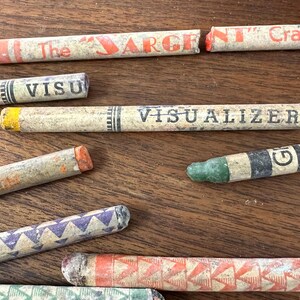 May include: A collection of vintage crayons in various colors and designs, scattered on a wooden surface. Some crayons have text that reads "The Sargent Crayon", "Visualizer", and "City of New York". The crayons are in various states of wear.