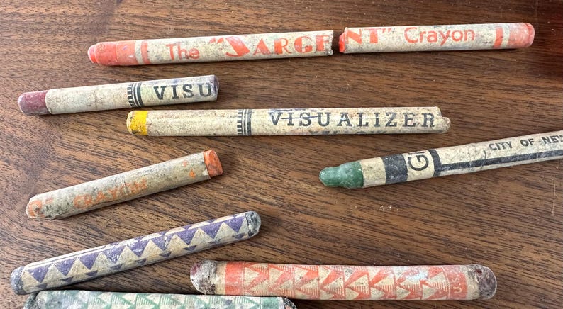 May include: A collection of vintage crayons in various colors and designs, scattered on a wooden surface. Some crayons have text that reads "The Sargent Crayon", "Visualizer", and "City of New York". The crayons are in various states of wear.