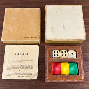 May include: A vintage Longchamp dice game set. The set includes three dice, colorful chips, and a leather case. The box is tan with the Longchamp logo. The game also includes a paper with the text "LE 421".