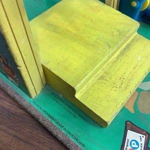 K&ouml;nnte beinhalten: Gelbes Holzspielzeug mit gr&uuml;nem Sockel und blau-gelbem Oberteil. Das Spielzeug hat ein musikalisches Spielwerk, das in der Schweiz hergestellt wurde. Der Text auf dem Sockel lautet "Pat. No. 3,114,216 Other Pats. Pending Patented Canada 1964 Musical Movement Made in Switzerland."