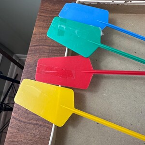 Puede incluir: Cuatro paletas de pl&aacute;stico de colores azul, verde, rojo y amarillo. Cada paleta tiene una cabeza rectangular y un mango largo y delgado. Las paletas est&aacute;n dispuestas en una caja sobre una superficie de madera.