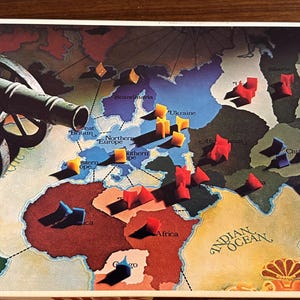 May include: A vintage Risk board game box with a red and white "RISK" logo. The box features a map of the world with colorful plastic army pieces and a cannon in the foreground.
