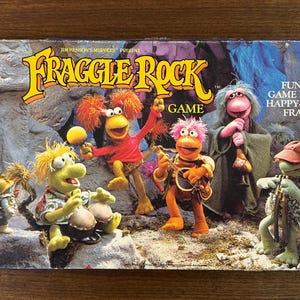 May include: Vintage Fraggle Rock board game box featuring colorful characters from the show. The box includes the game title, age range, and the Milton Bradley Company logo. The box is on a wooden surface.