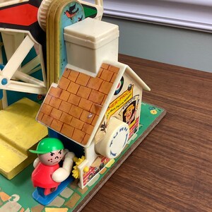 K&ouml;nnte beinhalten: Vintage Fisher-Price Little People Spielzeug-Riesenrad mit einem roten, gelben und blauen Rad, einem wei&szlig;en Haus mit braunem Dach und einer gr&uuml;nen und roten Little People Figur. Das Spielzeug hat eine Kurbel an der Seite, auf der "Drehen" und "Tickets" steht.