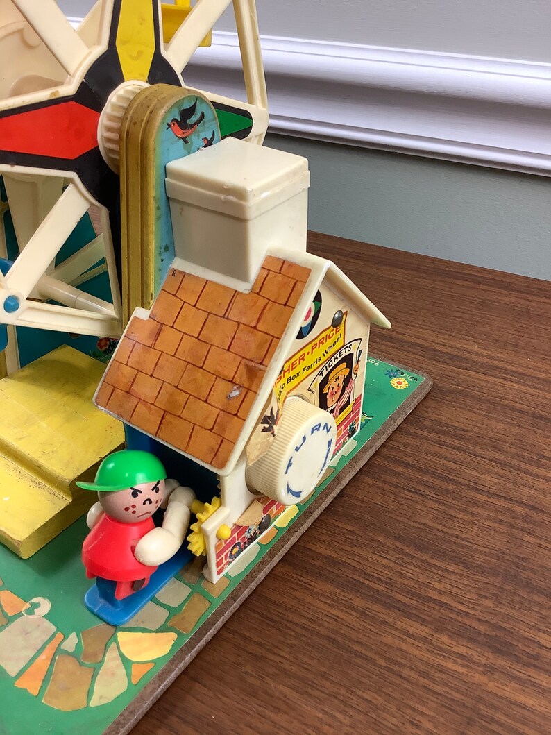 K&ouml;nnte beinhalten: Vintage Fisher-Price Little People Spielzeug-Riesenrad mit einem roten, gelben und blauen Rad, einem wei&szlig;en Haus mit braunem Dach und einer gr&uuml;nen und roten Little People Figur. Das Spielzeug hat eine Kurbel an der Seite, auf der "Drehen" und "Tickets" steht.