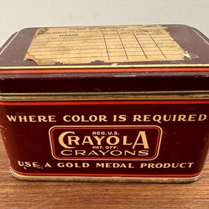 May include: A vintage Crayola crayons tin box. The rectangular box is dark red with gold and orange trim. The text "WHERE COLOR IS REQUIRED" and "CRAYOLA CRAYONS" are displayed on the front. The lid has a faded label.