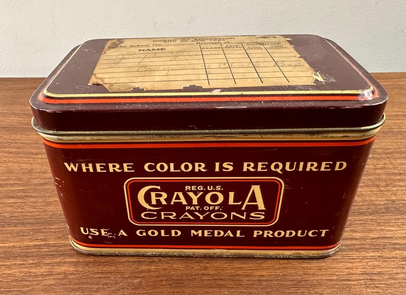 May include: A vintage Crayola crayons tin box. The rectangular box is dark red with gold and orange trim. The text "WHERE COLOR IS REQUIRED" and "CRAYOLA CRAYONS" are displayed on the front. The lid has a faded label.