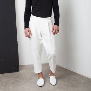 May include: A man wearing a black turtleneck sweater and white pants. He is standing on a white surface with a black background.