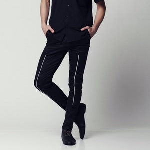 May include: A person wearing a black short-sleeved button-down shirt with white trim and black pants with white stripes. The person has silver hair and is standing with their hands in their pockets.