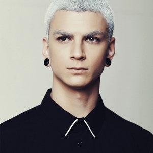 May include: A man with short, silver hair wearing a black collared shirt with white trim. He is wearing black earrings.