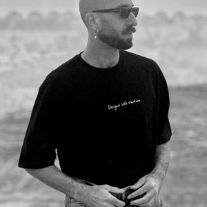 May include: A man wearing a black t-shirt with the text "Design with emotion" printed on it, sunglasses, and blue jeans. He is standing in a desert landscape with a cloudy sky in the background.