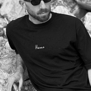 May include: A person wearing a black t-shirt with the word "Human" printed on it, sunglasses, and blue jeans. The person is sitting on a pile of rocks.