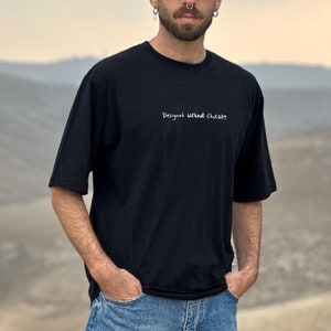 May include: A person wearing a black t-shirt with the text "Destroy What Creates" in white, blue jeans, and green sneakers. The person is standing on a rock in a desert landscape.