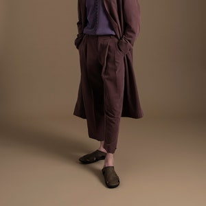 May include: A person wearing a long, burgundy coat, burgundy pants, and brown sandals. The person has short, dark hair and is standing on a brown background.