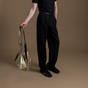 May include: A person wearing a black t-shirt with the word "HAMSÄ" printed on it, black pants, and black sandals. They are holding a gold-colored bag with a black strap.