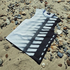 May include: A white sleeveless shirt with a black and white striped pattern. The shirt is lying on a sandy beach with small rocks.