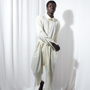 May include: Cream-colored jumpsuit with a collared shirt-style top and wide-leg pants. The outfit features a matching belt and button-down closure. The model is standing in front of a white backdrop.