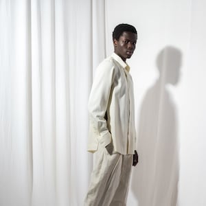 May include: A person wearing a white long-sleeved shirt and beige pants. They are standing in front of a white backdrop.