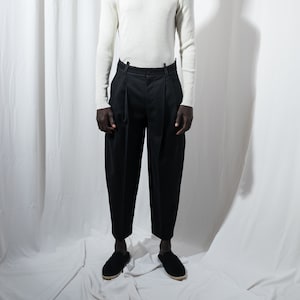 May include: A person wearing a white turtleneck sweater and black pants. The person is standing in front of a white background.