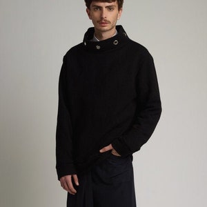 May include: A black mock neck sweater with a high collar and silver grommets. The sweater is paired with dark blue pants and black shoes.