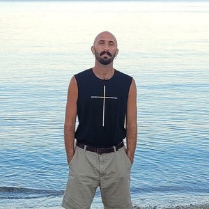 May include: A man wearing a black sleeveless shirt with a gold cross design, khaki shorts, and black sneakers. He is standing on a beach with a body of water in the background.