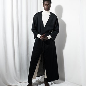 May include: A person wearing a long black robe with a white turtleneck underneath. The robe has a long sash that is tied at the waist. The person is standing on a white surface.