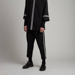 May include: A black long-sleeved shirt with white stripes on the sleeves and down the front. The shirt is paired with black pants that also have white stripes down the sides. The model is wearing black shoes.