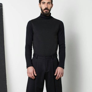 May include: A man wearing a black turtleneck sweater, black shorts, and black shoes. He is standing in front of a white wall.