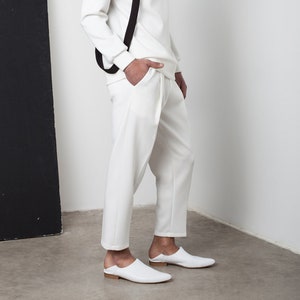 May include: A white turtleneck sweater with a black harness and white pants. The person is wearing white leather loafers.