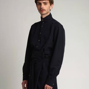 Puede incluir: Una camisa de manga larga azul marino oscuro con cuello alto y abotonadura. La camisa presenta un detalle envolvente en la cintura y pu&ntilde;os abotonados. La prenda est&aacute; hecha de una tela lisa y de color s&oacute;lido.