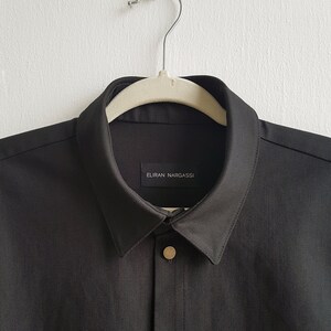 May include: A black collared shirt with a gold button and a label that reads "ELIRAN NARGASSI".