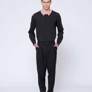 Puede incluir: Un hombre con una camisa negra de manga larga con cuello abotonado y pantalones negros. Est&aacute; de pie con las manos en los bolsillos y lleva zapatos negros.