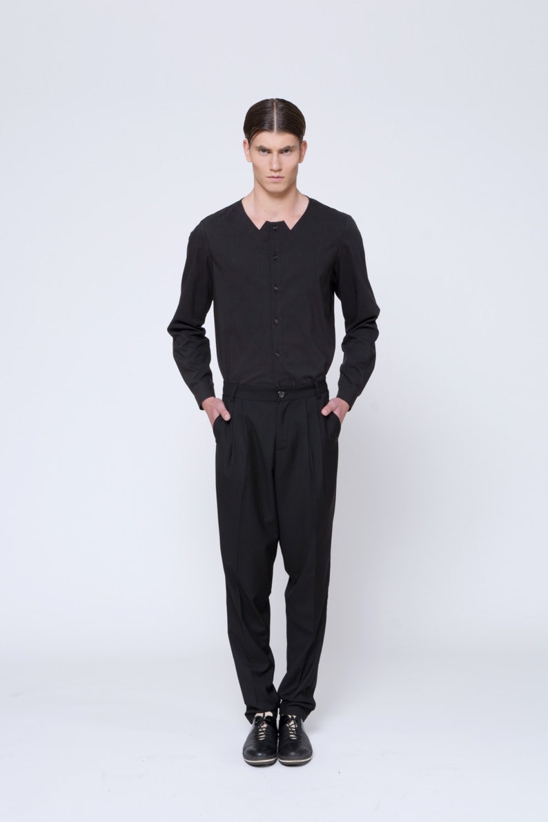 Puede incluir: Un hombre con una camisa negra de manga larga con cuello abotonado y pantalones negros. Est&aacute; de pie con las manos en los bolsillos y lleva zapatos negros.