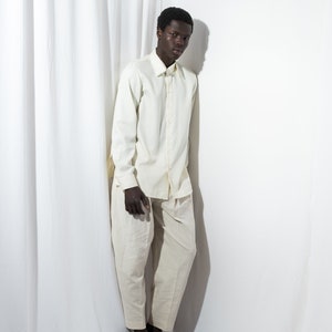 May include: A person wearing a white long-sleeved shirt and white pants. The person is standing in front of a white background.