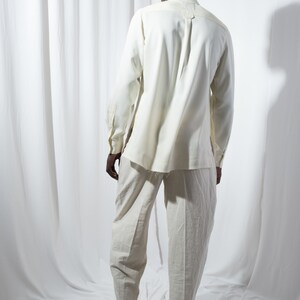 May include: A person wearing a white long-sleeved shirt and white pants. The person is standing in front of a white background.