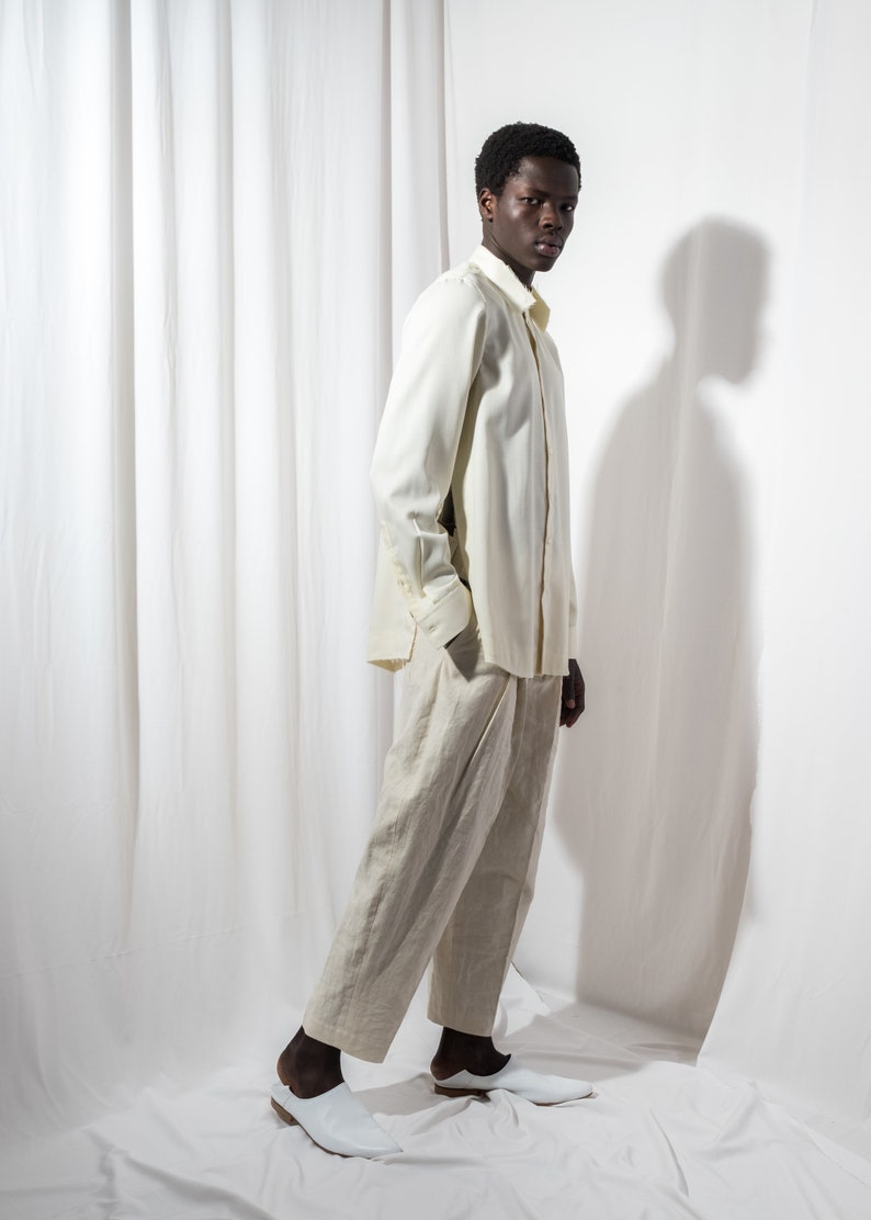 May include: A person wearing a white long-sleeved shirt and beige pants. They are standing in front of a white backdrop.