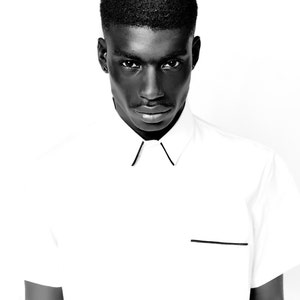 May include: A black and white photo of a person wearing a white collared shirt with black trim. The person has short black hair and a serious expression.