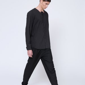 Puede incluir: Un hombre con una camisa negra de manga larga y pantalones negros. Est&aacute; caminando y mirando hacia abajo. Lleva zapatos negros.