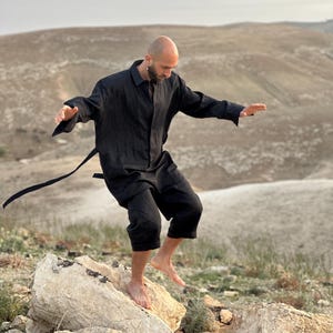May include: A person wearing a black linen shirt and pants stands on a rock formation. The shirt has a long, flowing silhouette and a relaxed fit. The person is barefoot and has a relaxed expression.