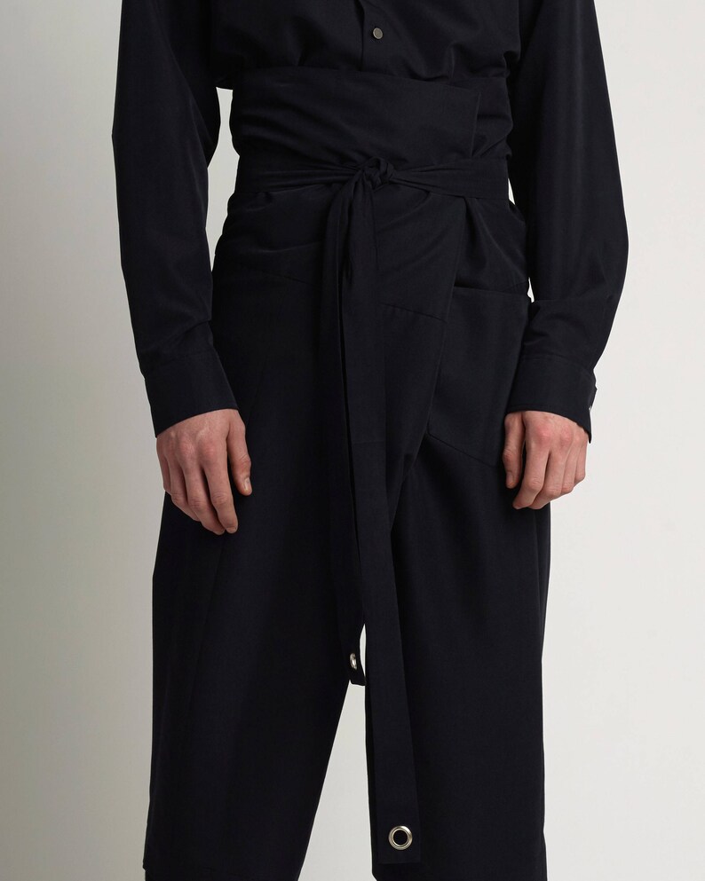Puede incluir: Conjunto negro que presenta una camisa de manga larga y pantalones de cintura alta con cintur&oacute;n envolvente. Los pantalones tienen un dise&ntilde;o &uacute;nico con cierre de lazo y ojales met&aacute;licos. El atuendo est&aacute; hecho de una tela oscura de color s&oacute;lido.