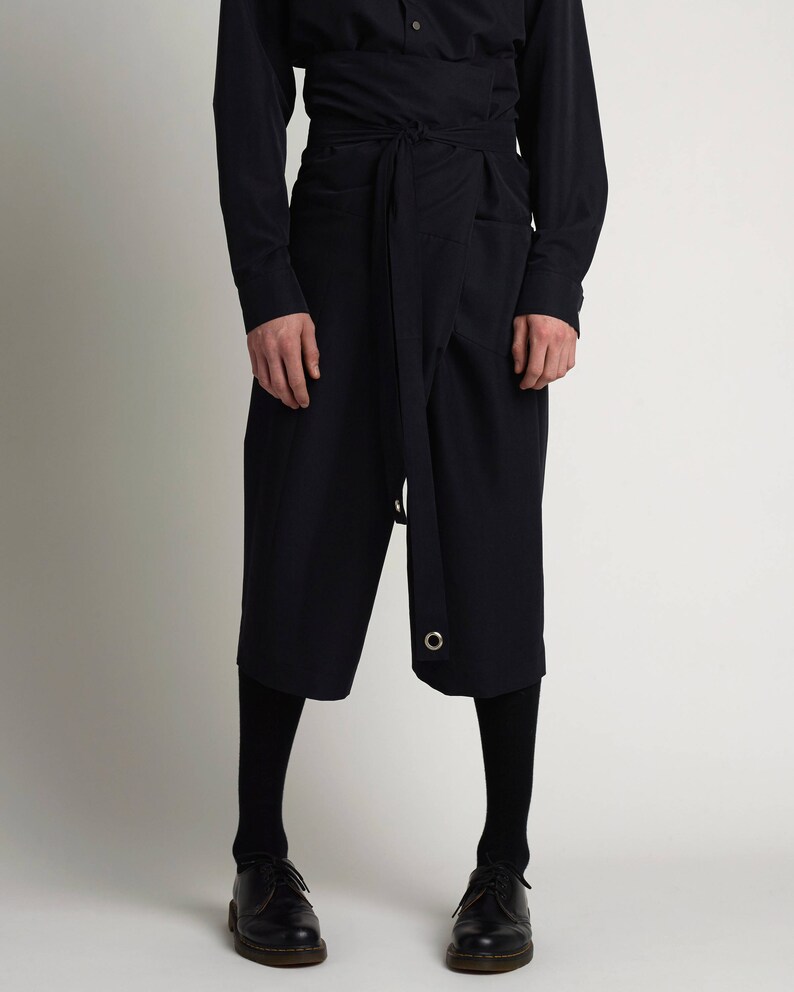 Puede incluir: Un conjunto azul marino oscuro con una camisa de manga larga y pantalones cortos tipo envolvente. Los pantalones cortos tienen un cintur&oacute;n que se envuelve alrededor de la cintura y un detalle de ojal met&aacute;lico. El atuendo se combina con medias negras y zapatos de cuero negros.