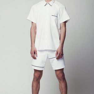 May include: A white short-sleeved button-down shirt with black trim and a white pair of shorts with black trim. The person is wearing black and white striped slip-on shoes.