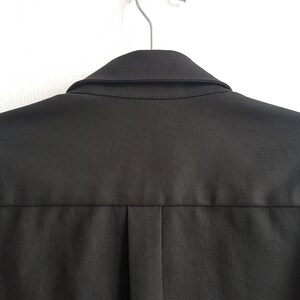 May include: A black collared shirt hanging on a silver hanger. The shirt has a single pleat in the back.