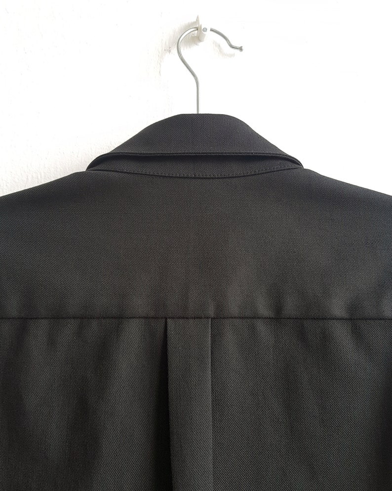 May include: A black collared shirt hanging on a silver hanger. The shirt has a single pleat in the back.