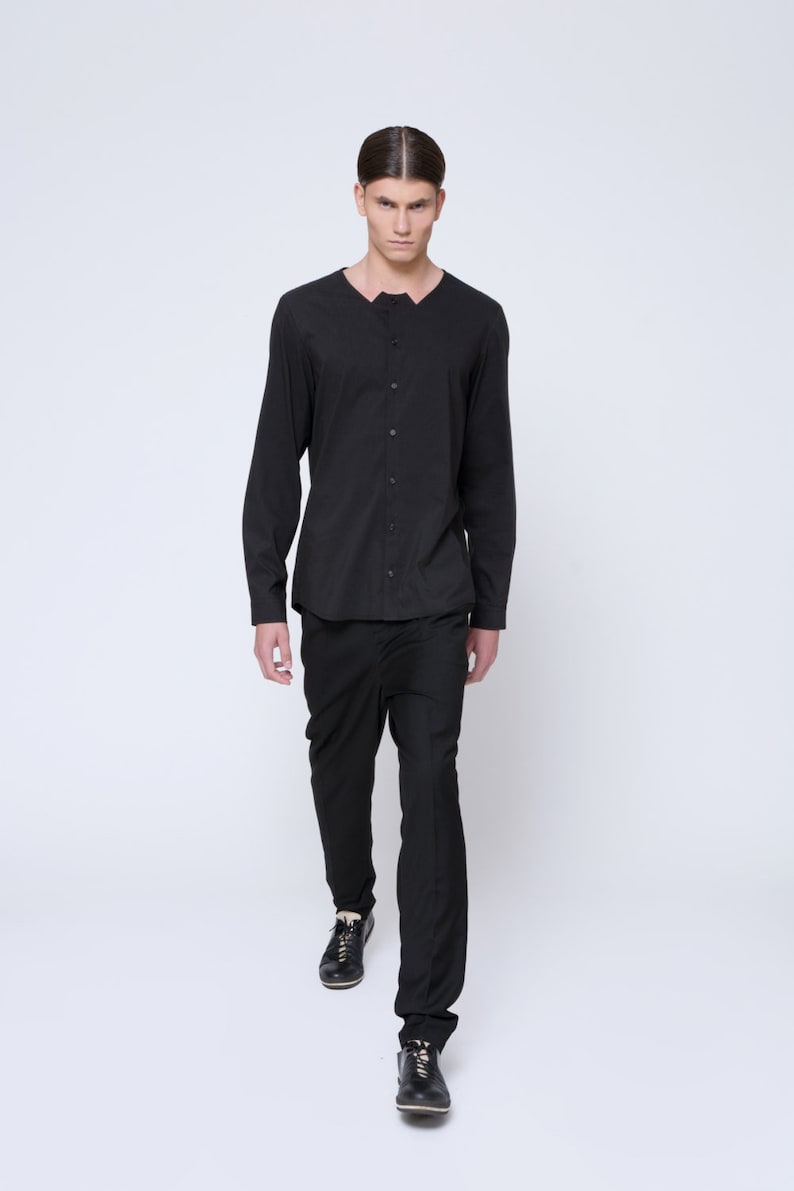 Puede incluir: Un hombre con una camisa negra de manga larga con cuello abotonado y pantalones negros. Lleva zapatos negros.