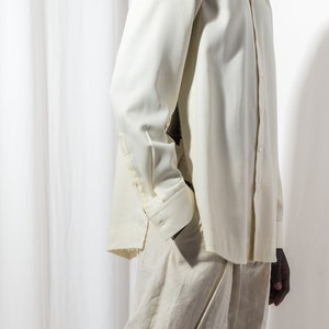 May include: A white long-sleeved shirt with a button-up collar and a white button-down shirt underneath. The shirt is tucked into white pants.