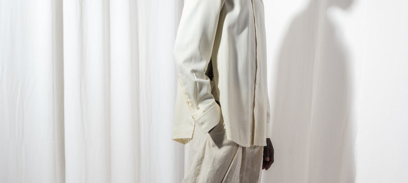 May include: A white long-sleeved shirt with a button-up collar and a white button-down shirt underneath. The shirt is tucked into white pants.
