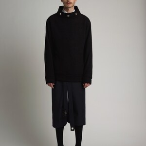 May include: A black mock neck sweater with a high collar and silver button accents. The sweater is paired with black pants and black shoes.
